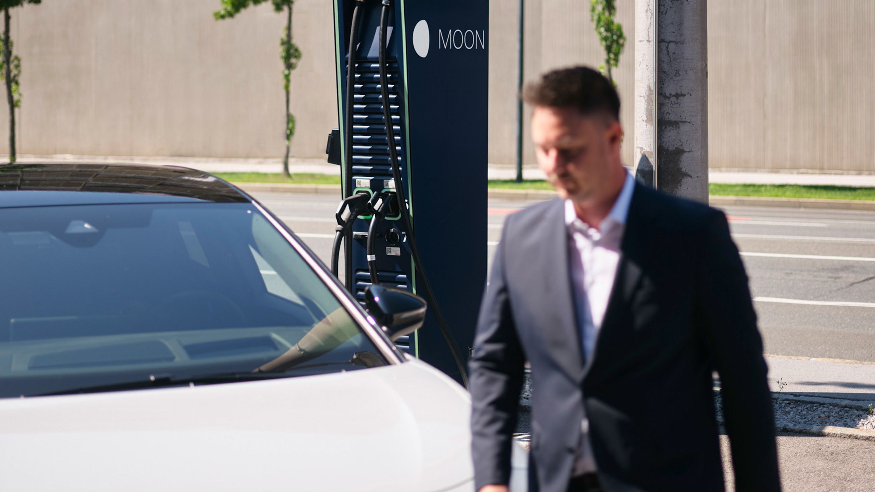 Moški hodi mimo električnega vozila pred hitro polnilno postajo.