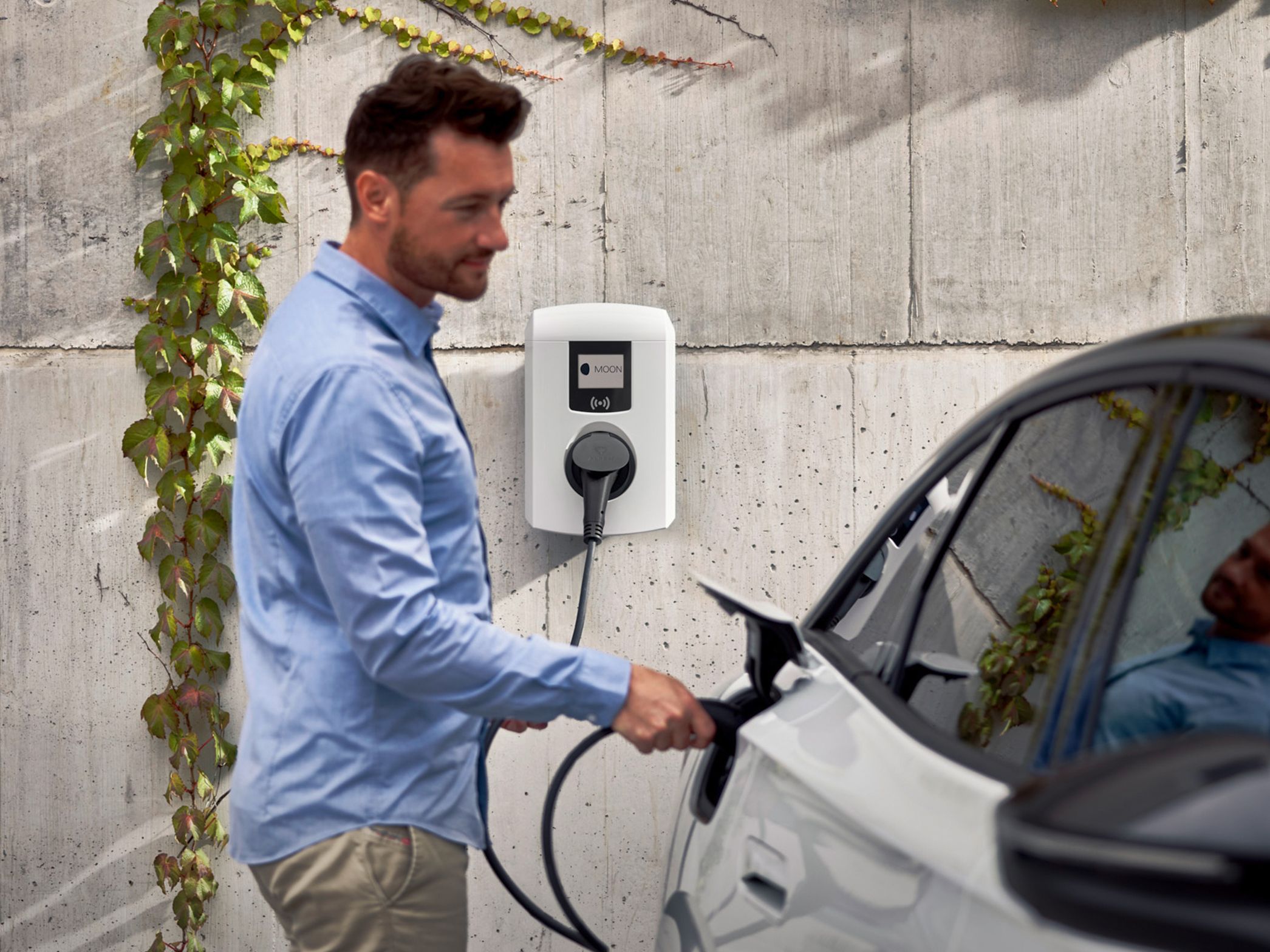 Moški pred stavbo priklaplja električni avto na stensko polnilnico.