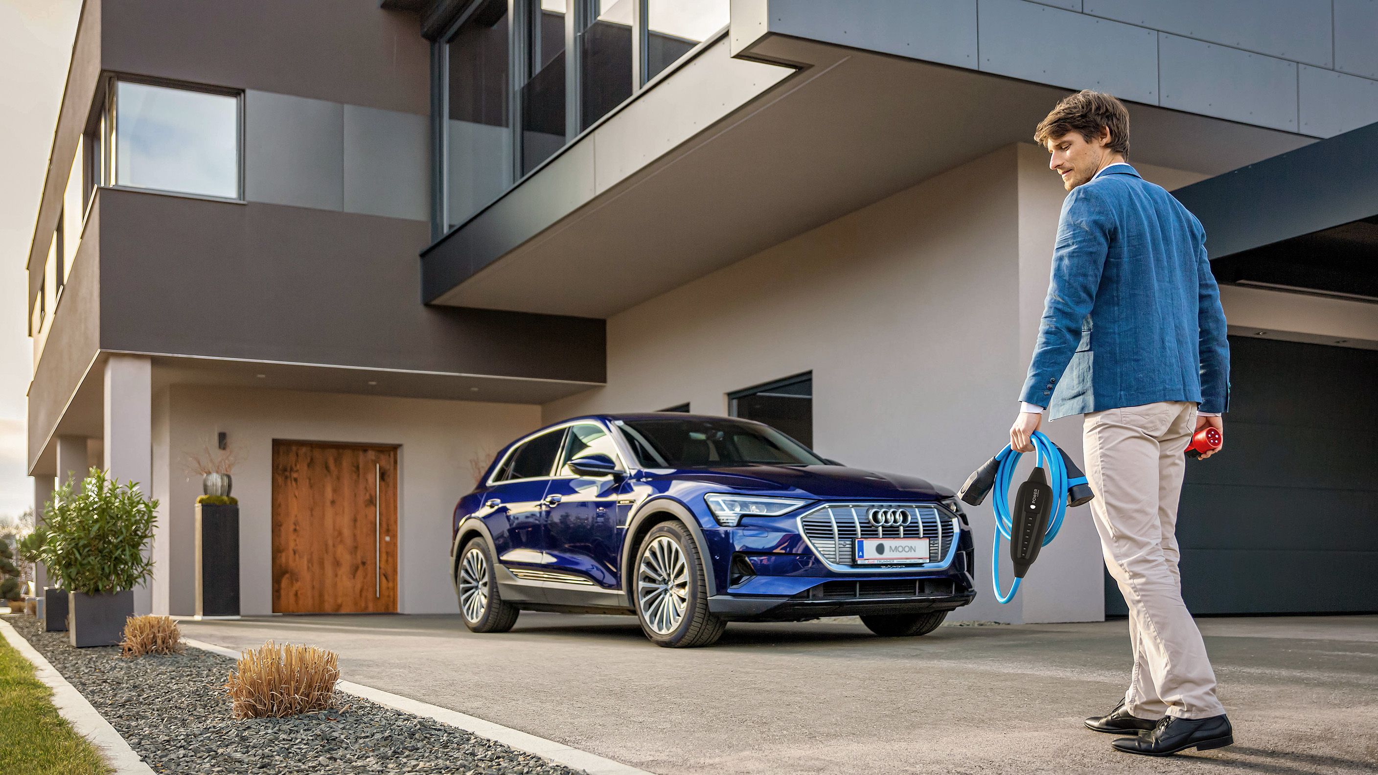 Moški se približuje električnemu avtomobilu ob hiši in v roki drži MOON POWER2Go polnilni kabel.