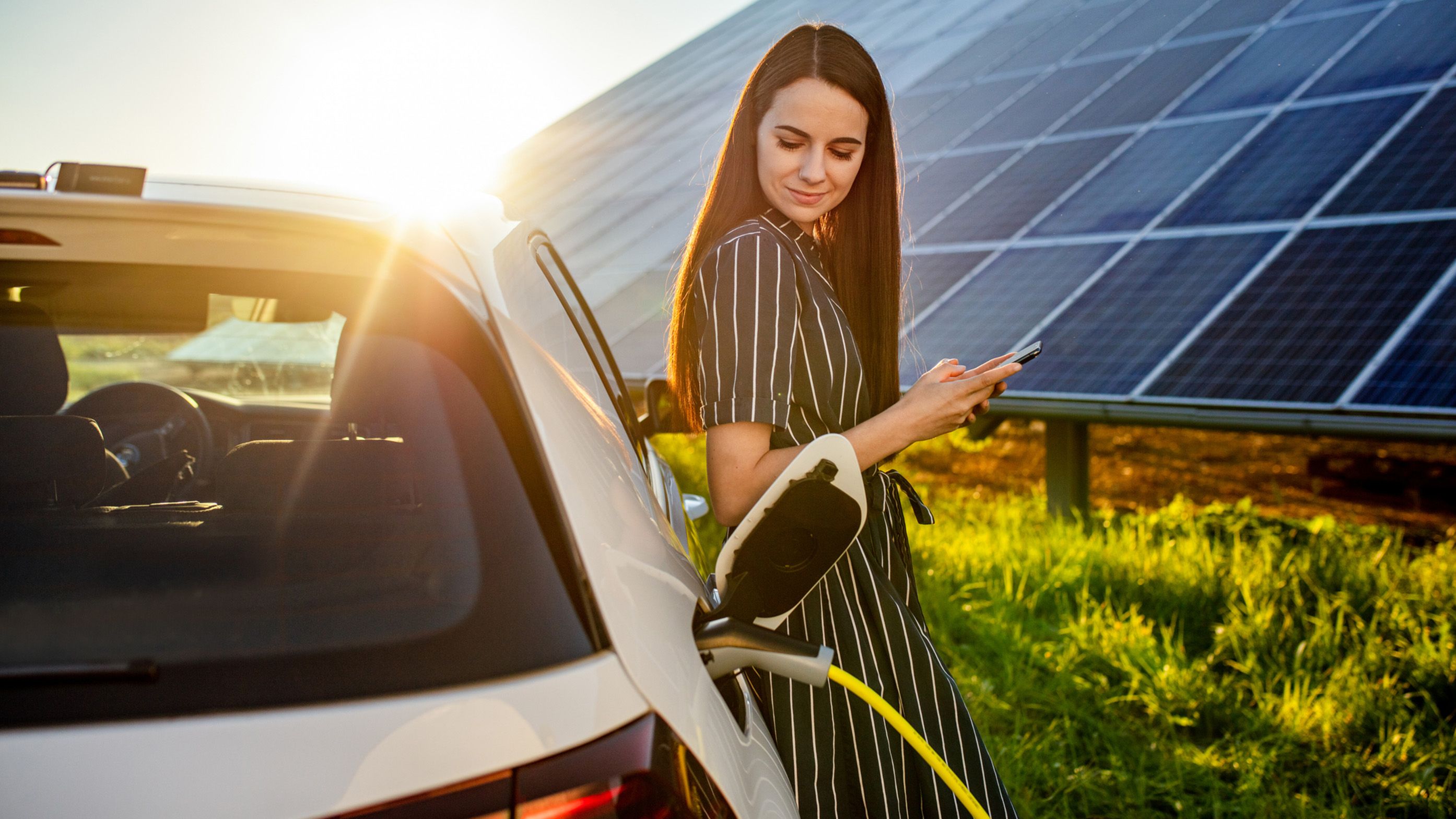 Ženska se naslanja na električni avtomobil, parkiran pred sončno elektrarno.