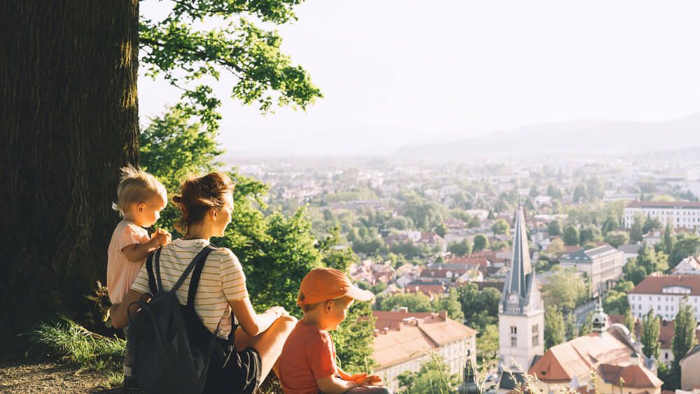 Mama z dvema majhnima otrokoma sedi na hribčku s pogledom na mesto.