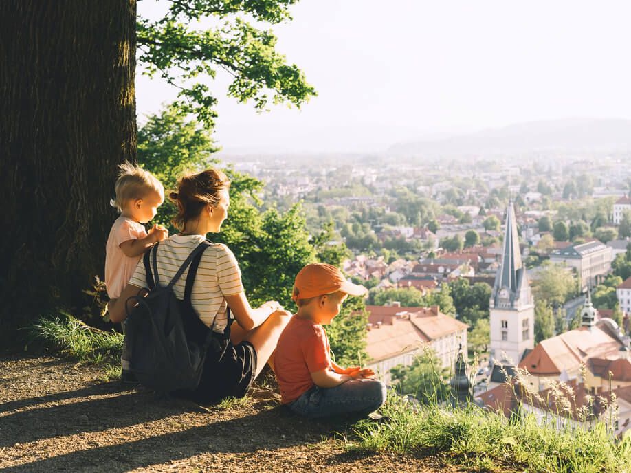 Mama z dvema majhnima otrokoma sedi na hribčku s pogledom na mesto.