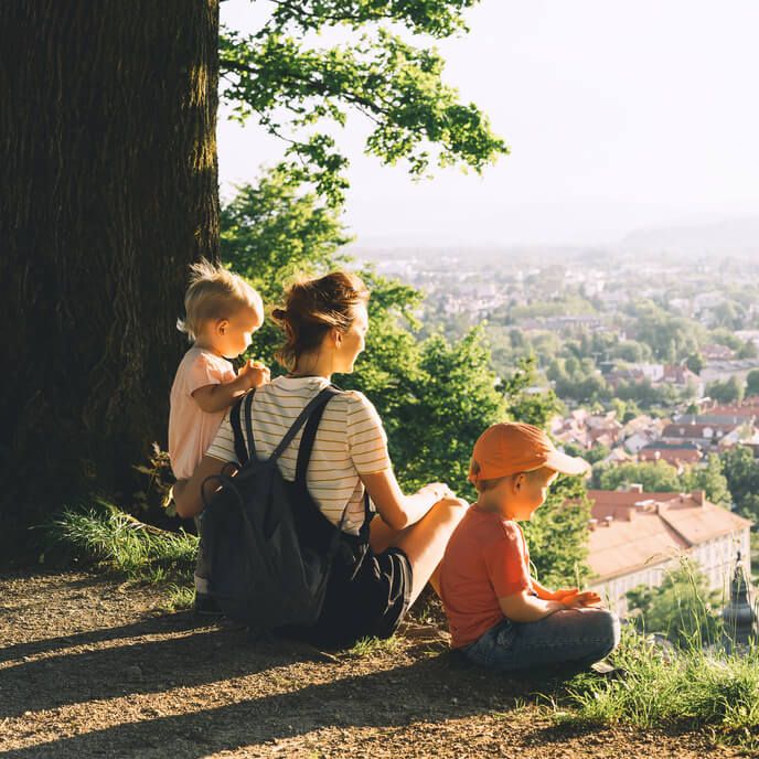 Mama z dvema majhnima otrokoma sedi na hribčku s pogledom na mesto.